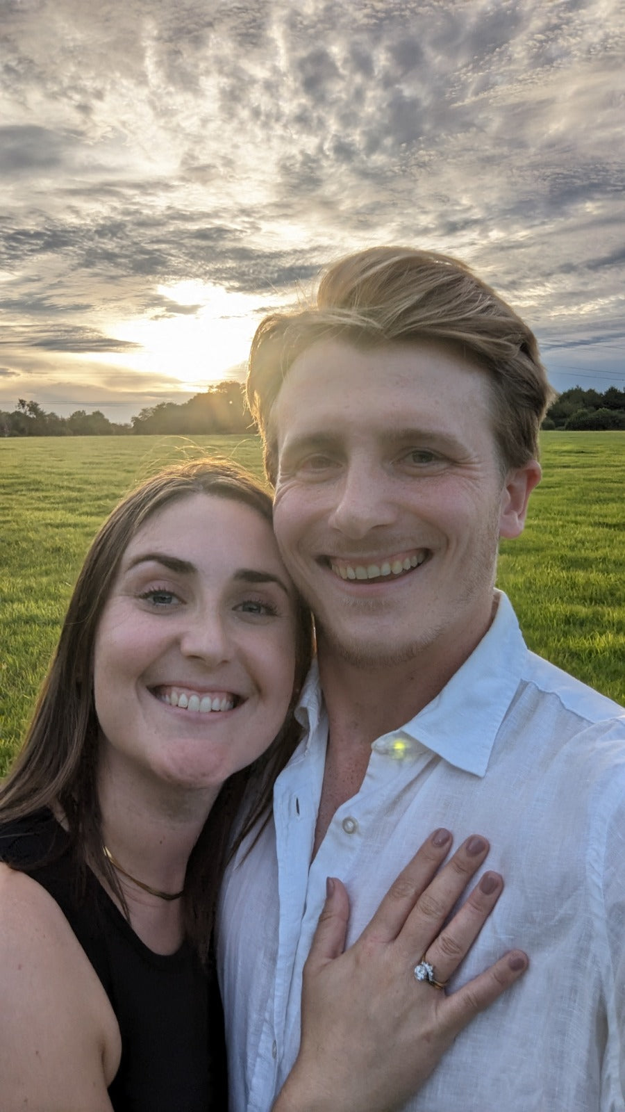Couple smiling outdoors in a field at sunset. Woman’s hand rests on man’s chest, displaying a visible engagement ring with a central gemstone - Sample Image