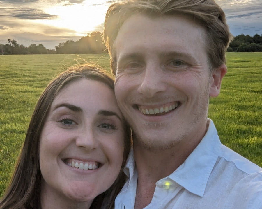 Couple smiling outdoors in a field at sunset. Woman’s hand rests on man’s chest, displaying a visible engagement ring with a central gemstone - Sample Image