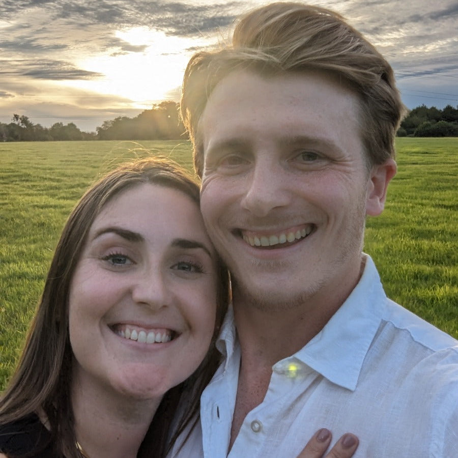 Couple smiling outdoors in a field at sunset. Woman’s hand rests on man’s chest, displaying a visible engagement ring with a central gemstone - Sample Image