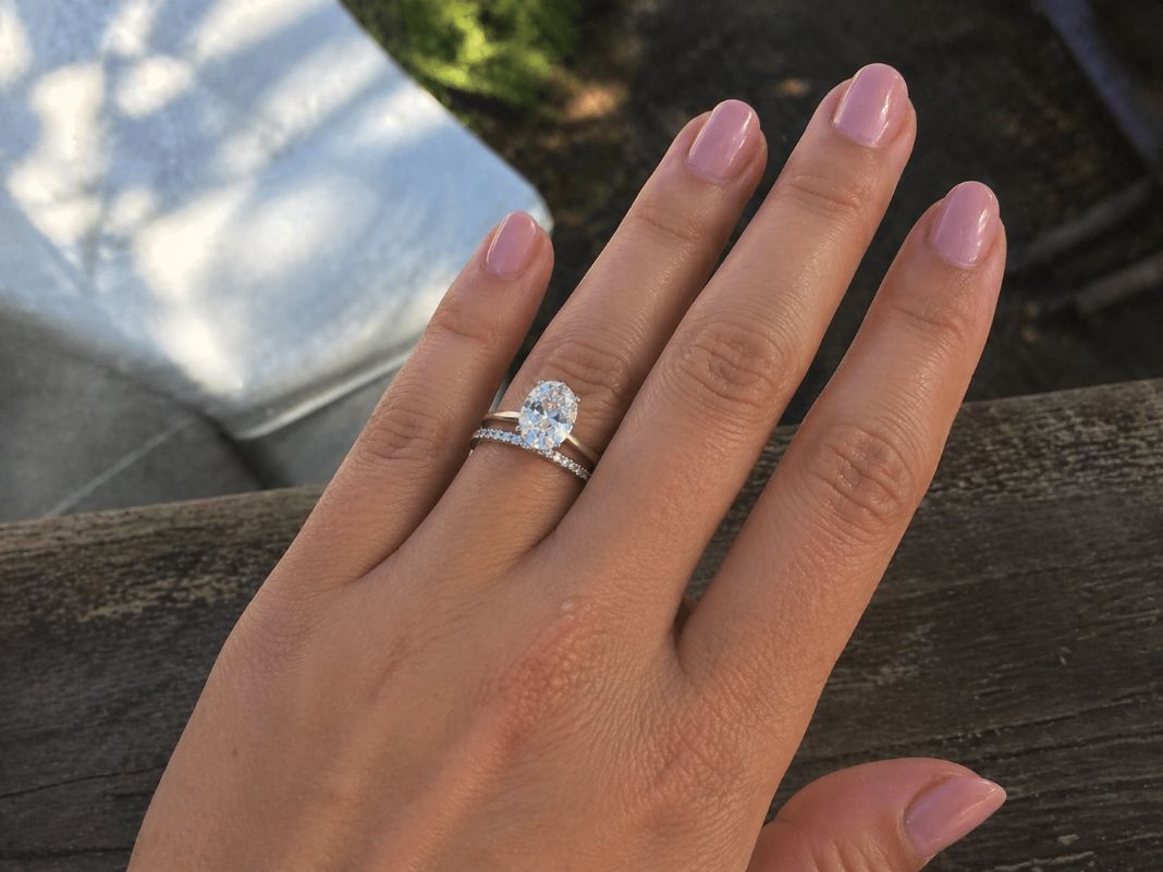 Engagement ring with an oval-cut diamond set on a white gold band, paired with a matching diamond ring guard, displayed on a hand outdoors. Elegant and timeless design, perfect for stacking or as a solitaire set. - Sample Image