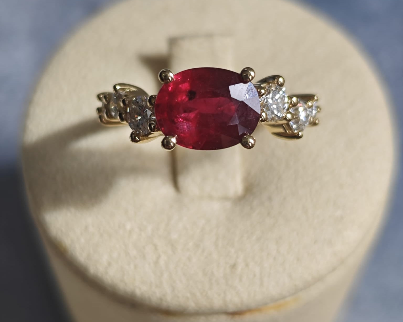 Gold ring with an oval red gemstone flanked by diamonds, displayed on a beige cushion. Elegant and vibrant design. - Sample Image