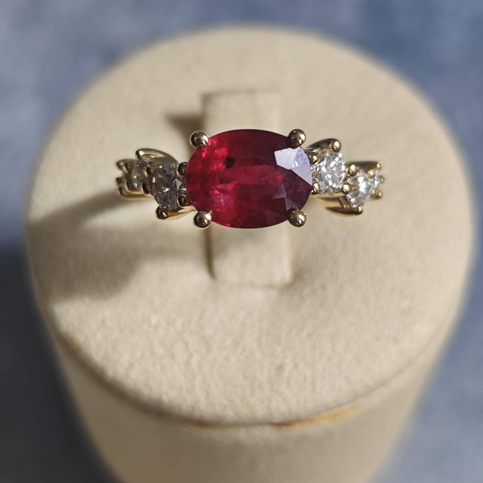 Gold ring with an oval red gemstone flanked by diamonds, displayed on a beige cushion. Elegant and vibrant design. - Sample Image