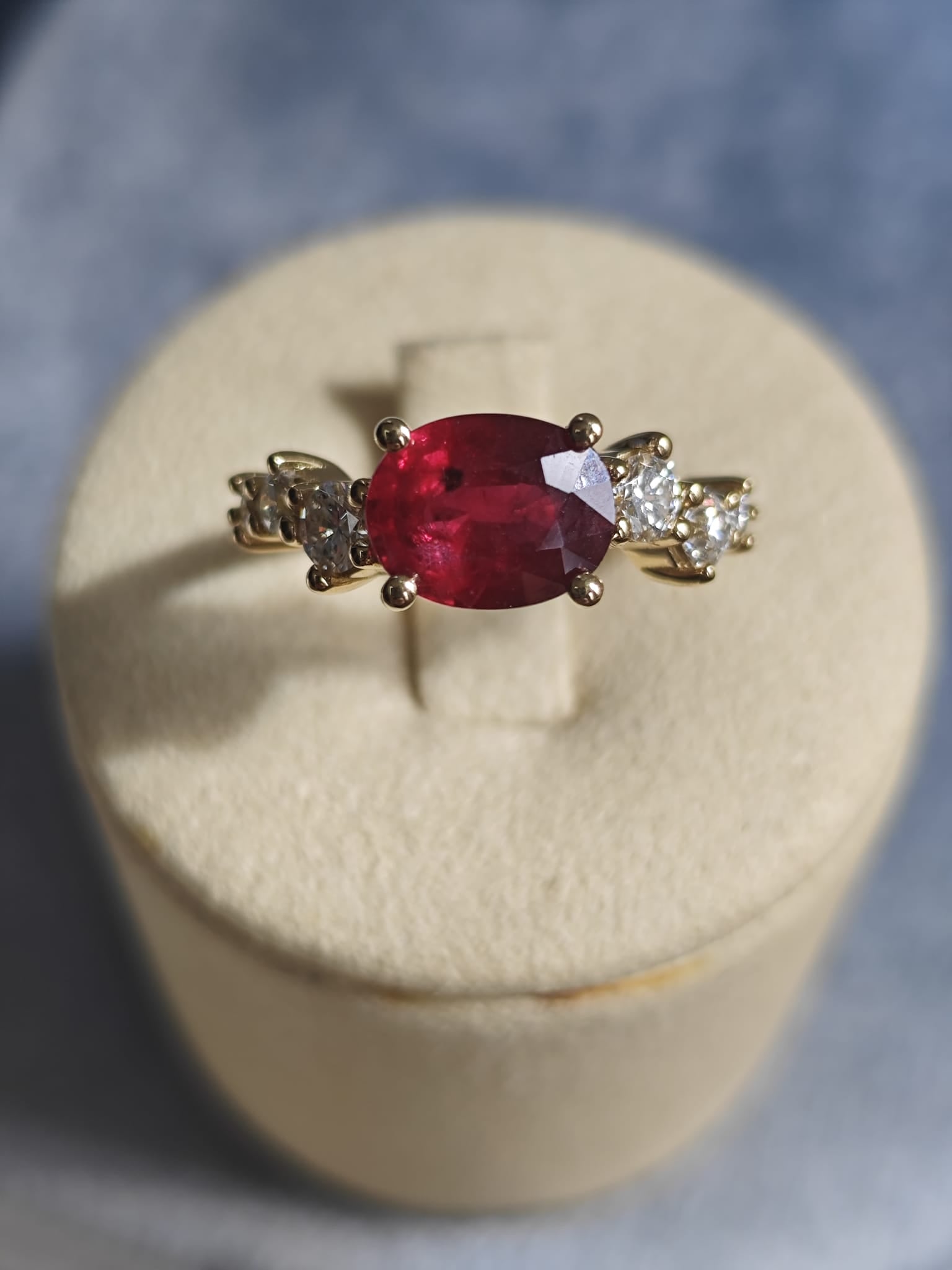 Gold ring with an oval red gemstone flanked by diamonds, displayed on a beige cushion. Elegant and vibrant design. - Sample Image
