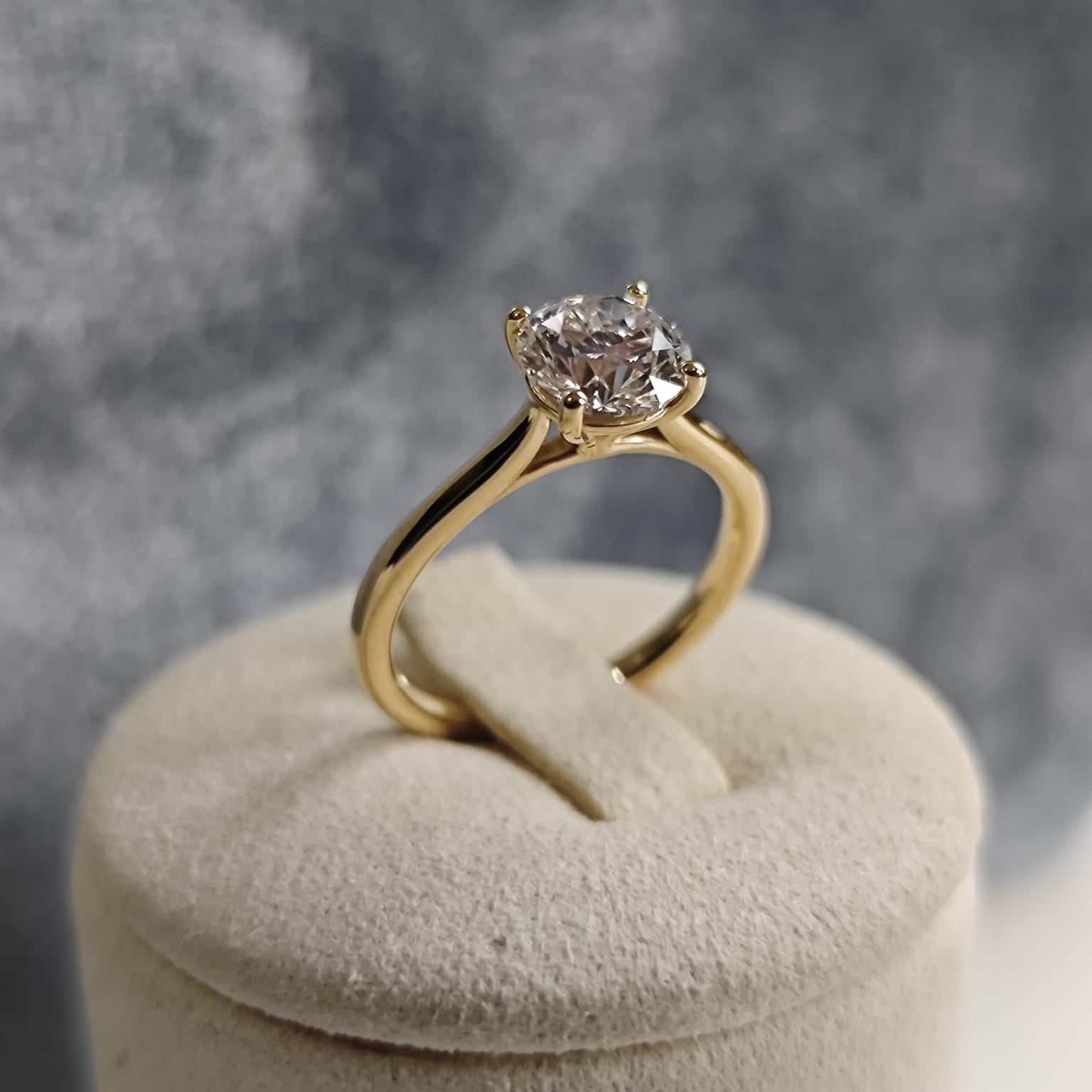 Gold solitaire ring with a round central gemstone, displayed on a beige cushion with a grey background. Classic and elegant design. - Sample Image