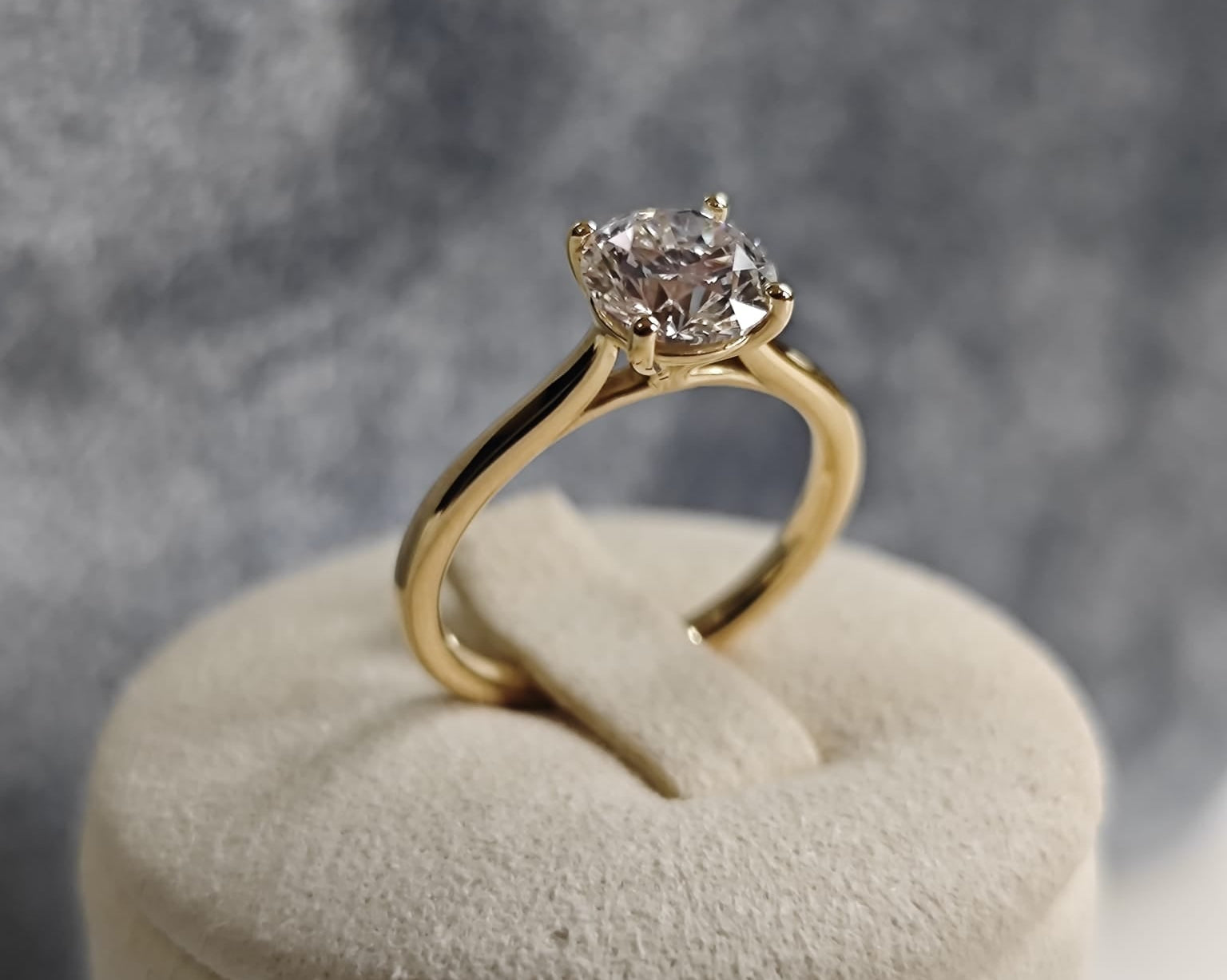 Gold solitaire ring with a round central gemstone, displayed on a beige cushion with a grey background. Classic and elegant design. - Sample Image