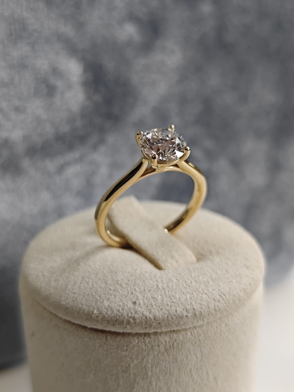 Gold solitaire ring with a round central gemstone, displayed on a beige cushion with a grey background. Classic and elegant design. - Sample Image