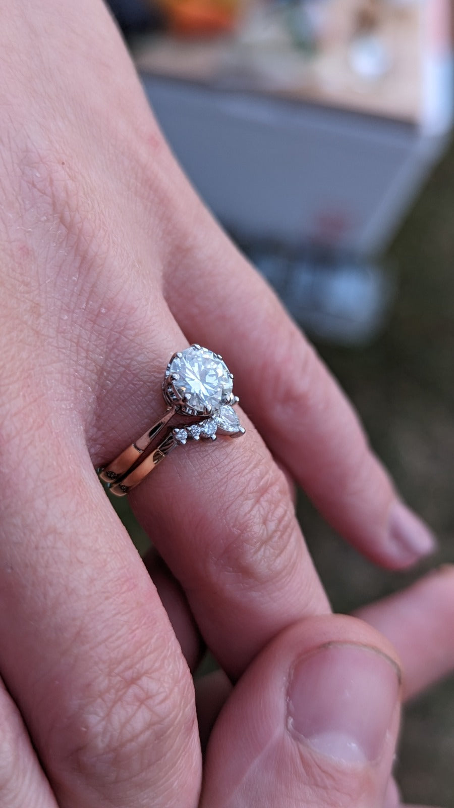 Hand wearing a two-ring set with a central round gemstone and smaller accent stones, displayed outdoors with a blurred background. Elegant engagement and wedding ring combination. - Sample Image