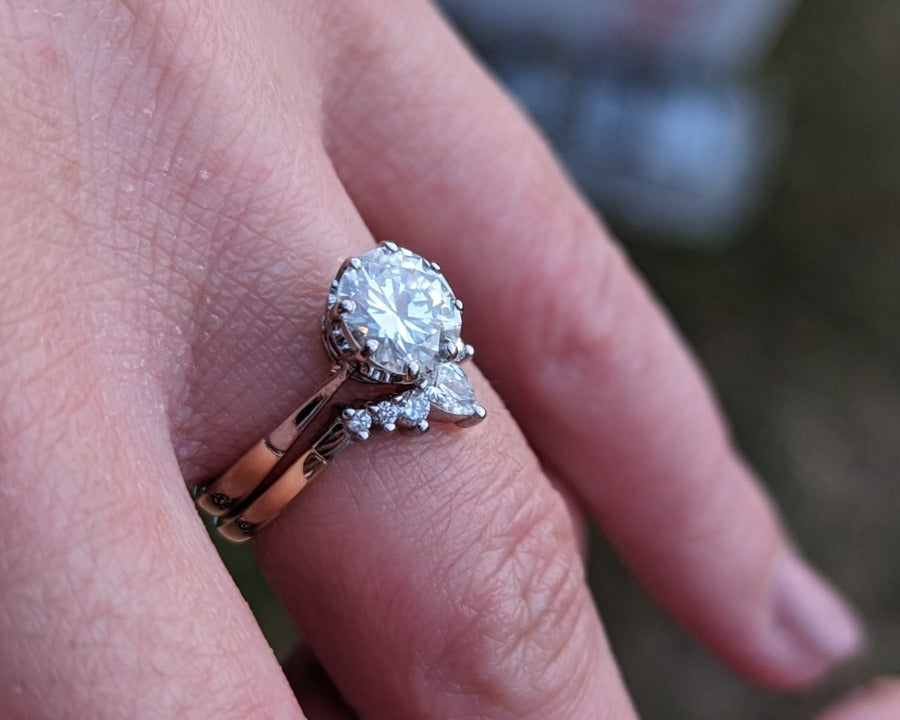 Hand wearing a two-ring set with a central round gemstone and smaller accent stones, displayed outdoors with a blurred background. Elegant engagement and wedding ring combination. - Sample Image