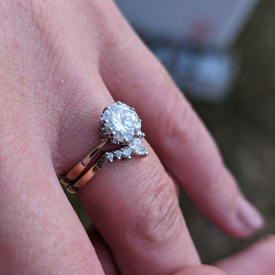 Hand wearing a two-ring set with a central round gemstone and smaller accent stones, displayed outdoors with a blurred background. Elegant engagement and wedding ring combination. - Sample Image