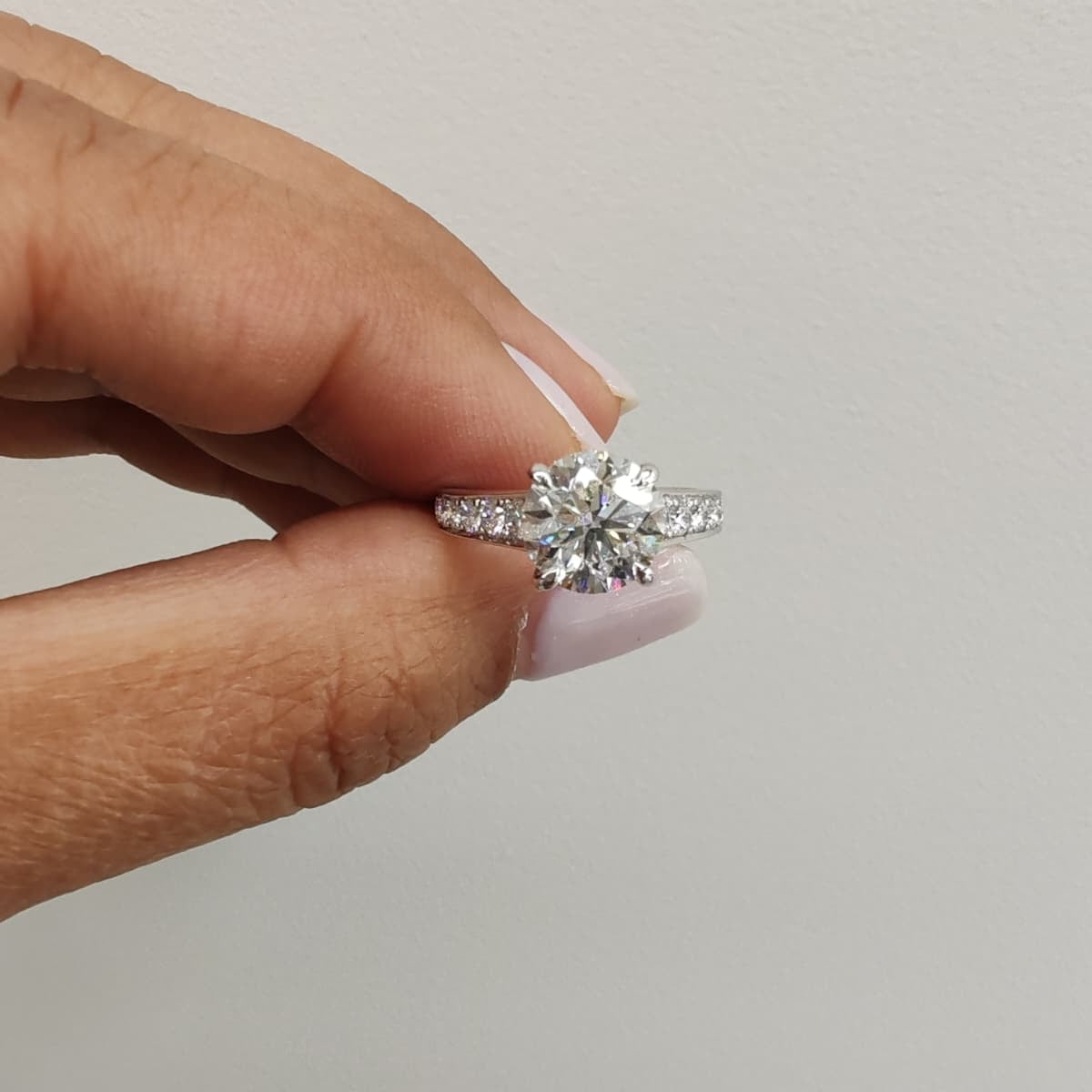 White gold engagement ring with a round brilliant-cut diamond, held between fingers against a plain background. Classic solitaire design with sparkling side stones for added elegance. - Sample Image