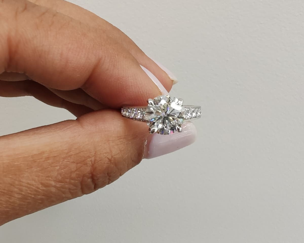 White gold engagement ring with a round brilliant-cut diamond, held between fingers against a plain background. Classic solitaire design with sparkling side stones for added elegance. - Sample Image