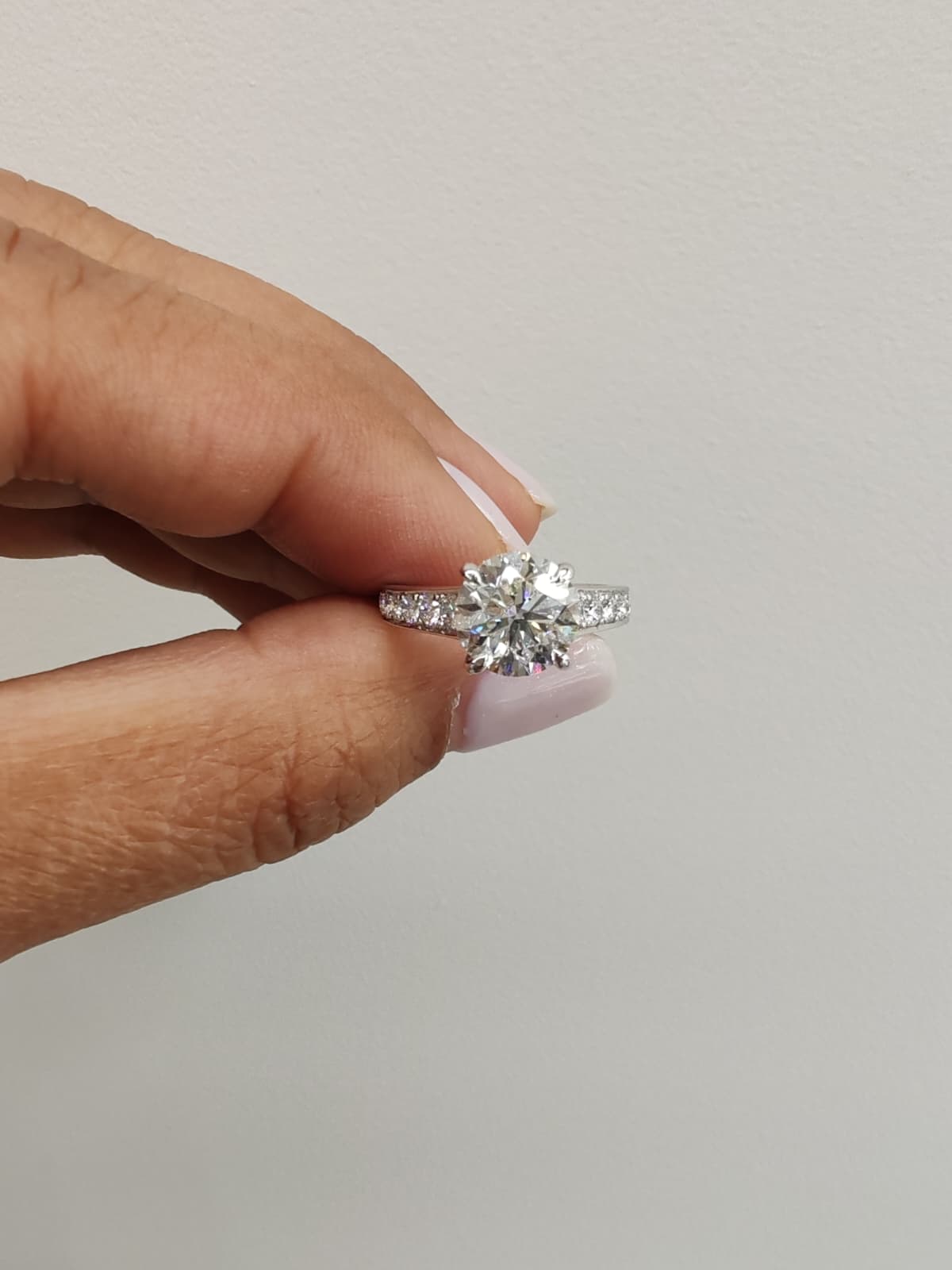 White gold engagement ring with a round brilliant-cut diamond, held between fingers against a plain background. Classic solitaire design with sparkling side stones for added elegance. - Sample Image