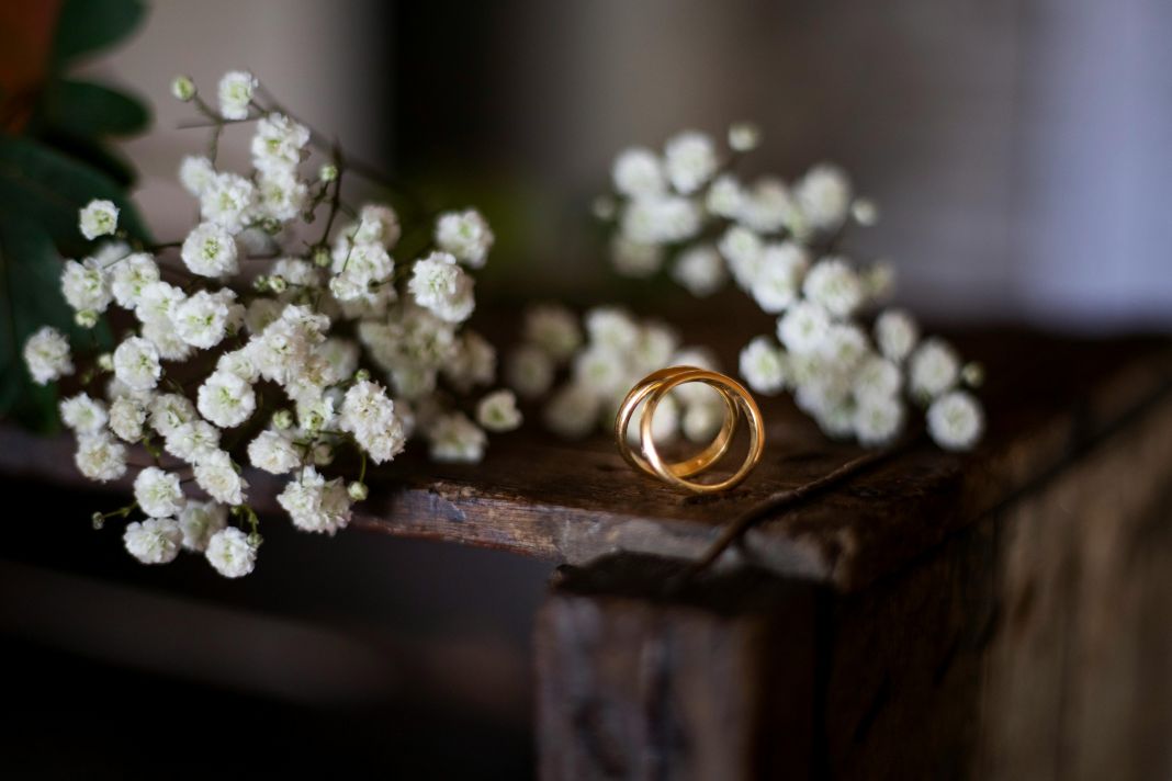 a photo of two gold wedding rings on a wooden desk with white flowers on the table behind them